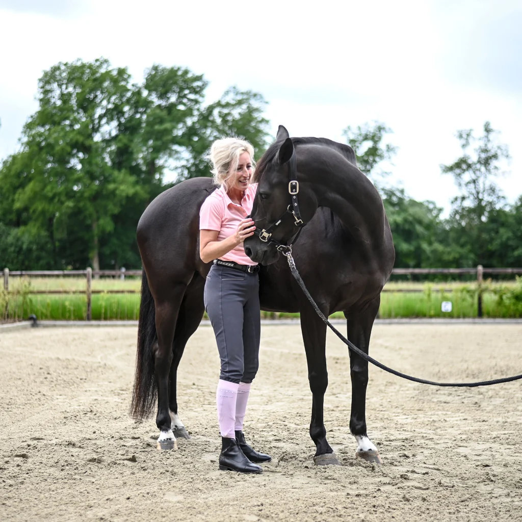 Sabrina Barmentloo van 'Osteopaat voor je Paard' staat lachend naast een zwart paard in een buitenomgeving. De afbeelding illustreert een blog over het gebruik van biotine supplementen voor de gezondheid van paardenhoeven, met uitleg over de werking, voordelen, en wetenschappelijk bewijs.